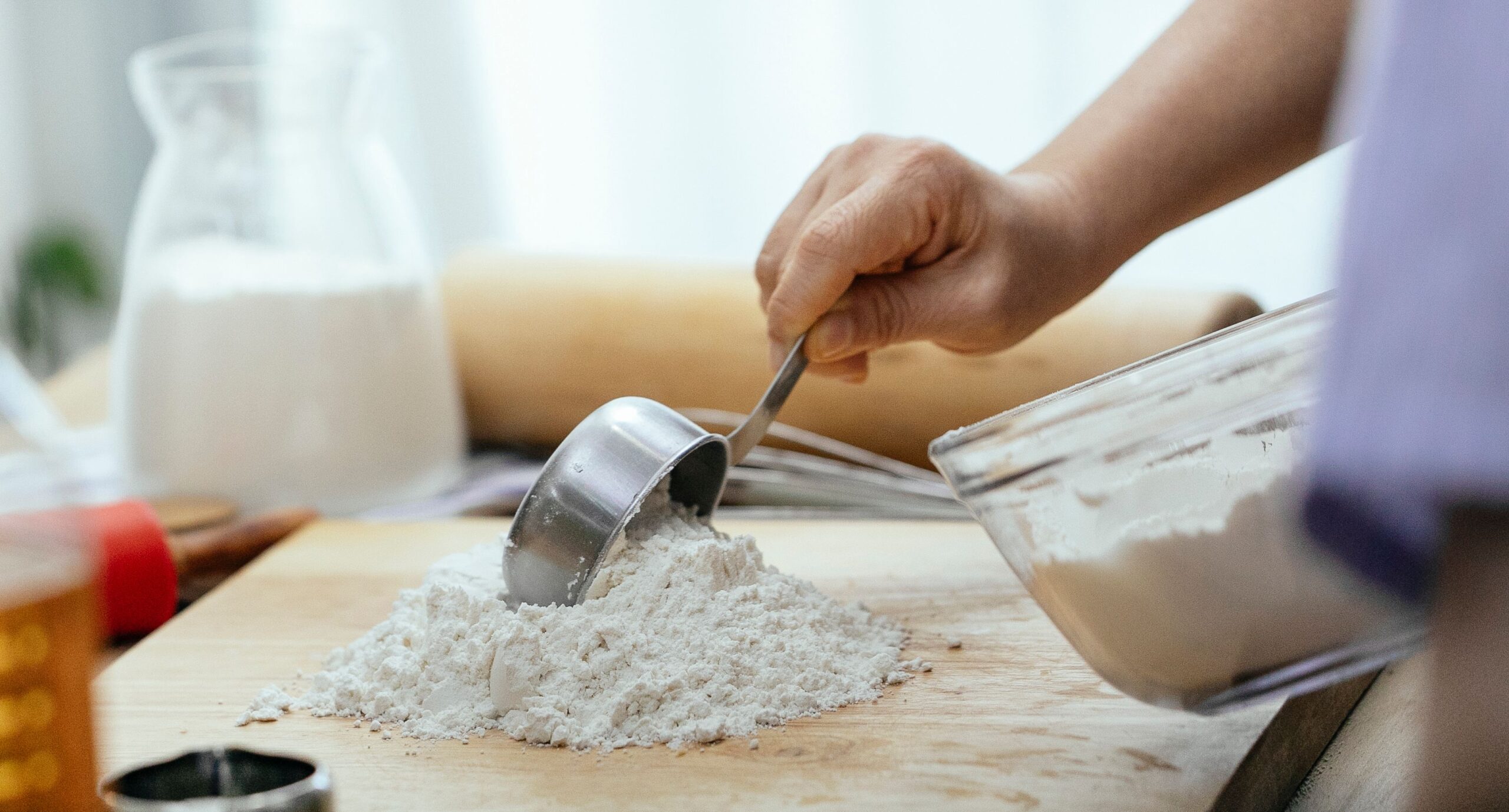 A hand spooning flour into a measuring cup with a straight-edged knife nearby for leveling.
